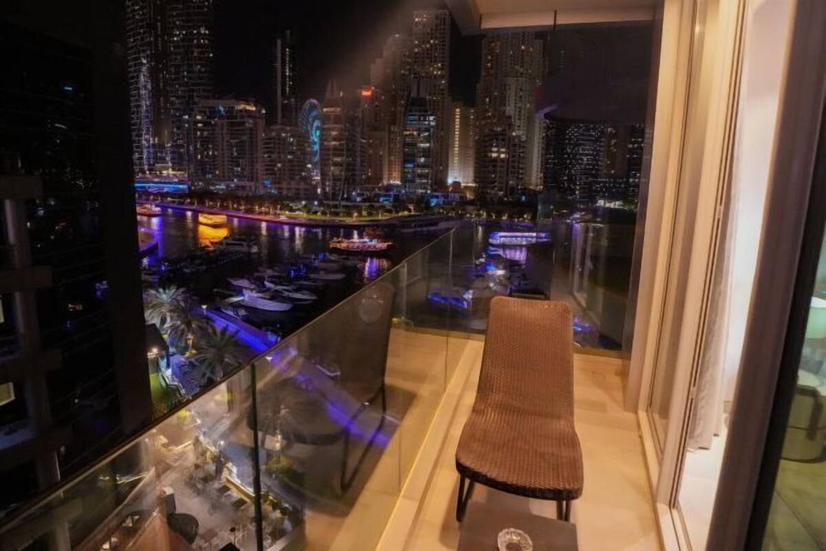 A balcony with a chair, a glass railing, and a cityscape view at night.
