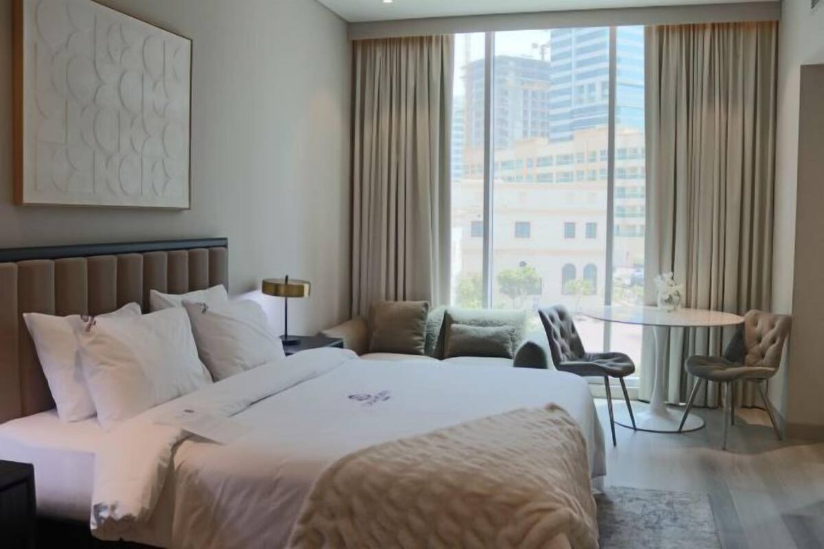 A hotel room with a large bed, a desk, a chair, and a view of buildings through the window.