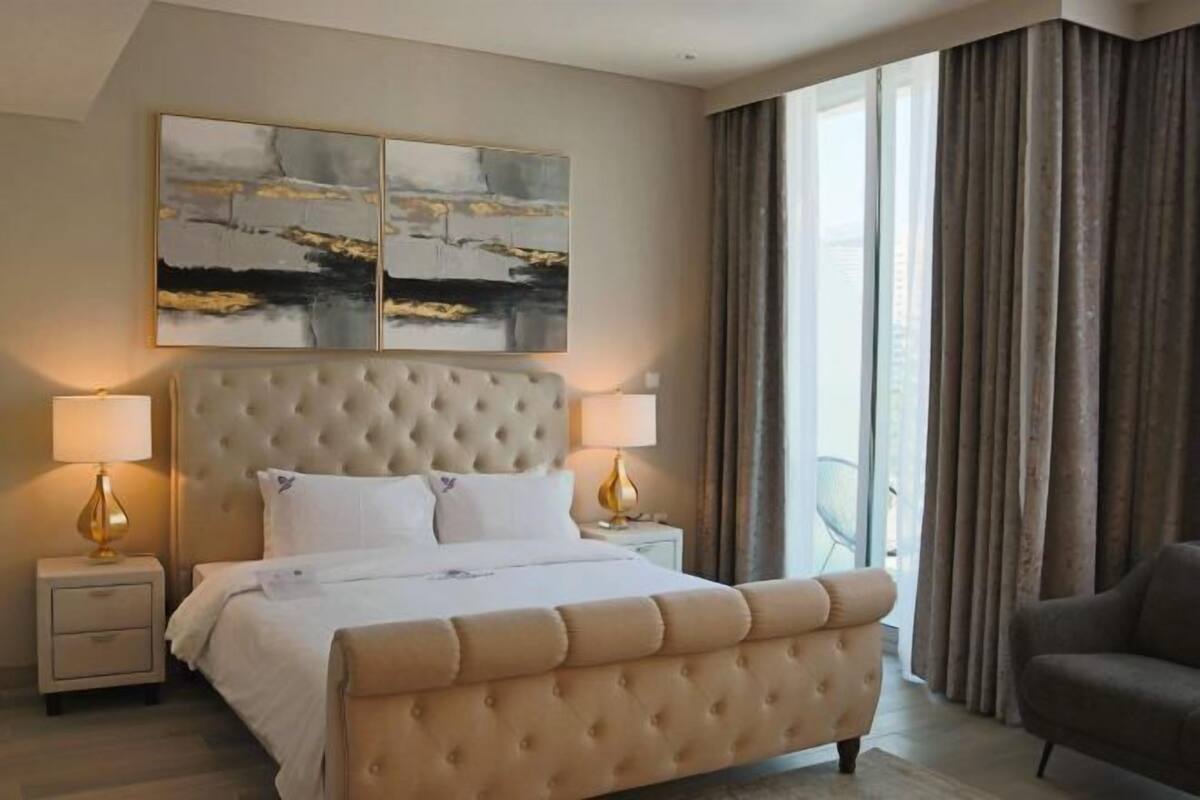 A hotel room with a large bed, two bedside tables, a headboard, and a view of the outdoors.