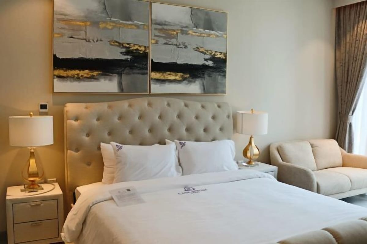 Una habitación de hotel con tablero de cabecera capitonado, cama con ropa de cama blanca, dos lámparas de noche, una silla y una obra de arte abstracta en la pared.
