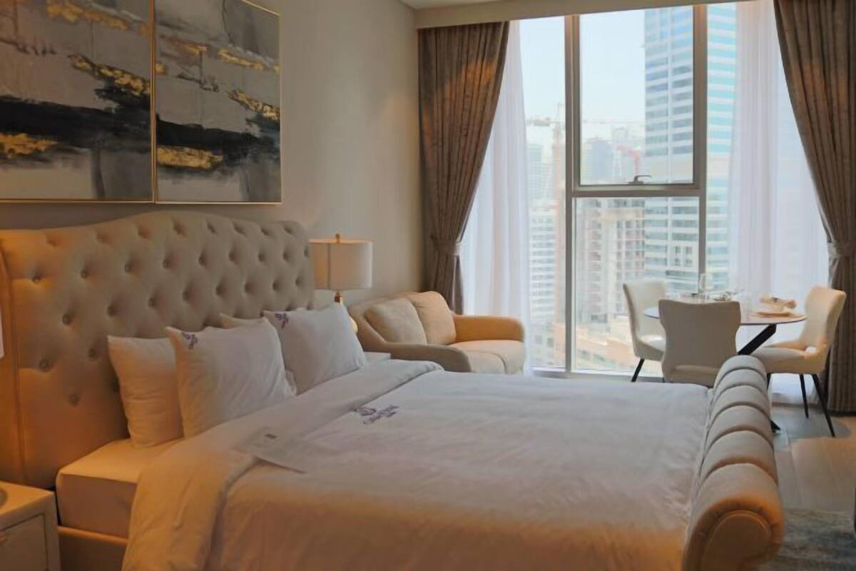 A hotel room with a large bed, a sitting area with a table and chairs, and a view of the city through large windows.