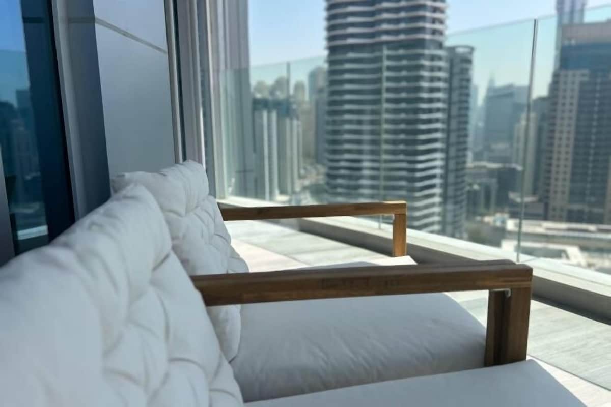 Una habitación moderna con un banco acolchado blanco junto a un ventanal grande, con vistas a edificios altos.