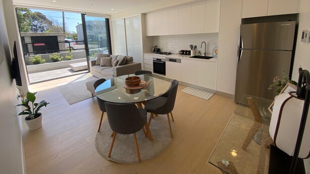 A modern kitchen with white cabinets, a stainless steel refrigerator, and a dining area with a glass table and chairs. - Executive Oasis | Chic 1BR + Courtyard Retreat (Engadine)