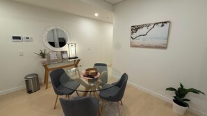 A modern dining area with a glass table, four grey chairs, a wooden side table with a vase and a framed certificate, a round mirror, a wall-mounted air conditioner, a trash bin, and a potted plant. - Executive Oasis | Chic 1BR + Courtyard Retreat (Engadine)