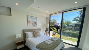A modern bedroom with a large bed, a bedside table with a lamp, a framed picture on the wall, and a view of the outside through a sliding glass door. - Executive Oasis | Chic 1BR + Courtyard Retreat (Engadine)