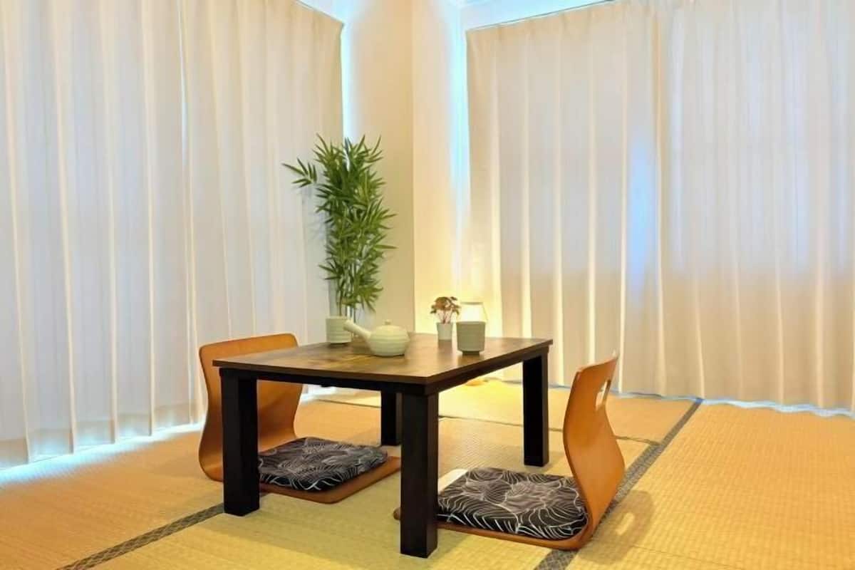 A traditional Japanese-style room with tatami flooring, a low wooden table, two cushioned chairs, a potted plant, and a lamp.