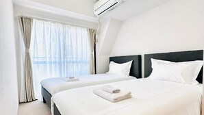 A hotel room with two beds, a large window with curtains, an air conditioning unit, and a neatly made bed with a towel. - City Heim Building (Tokyo)