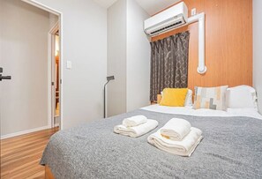 A hotel room with a bed, a grey blanket, two rolled towels, a wooden headboard, a grey wall, a white air conditioning unit, and a door leading to a hallway. - daiwa Hotel, Skytree, Ueno Park, Asakusa Temple (Tokyo)