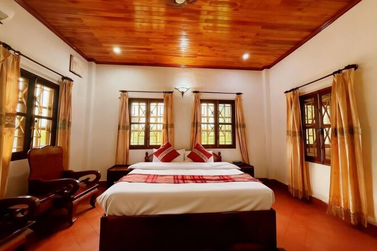 A bedroom with a wooden ceiling, two windows with curtains, a bed with a red and white checkered bedspread, and a wooden chair.