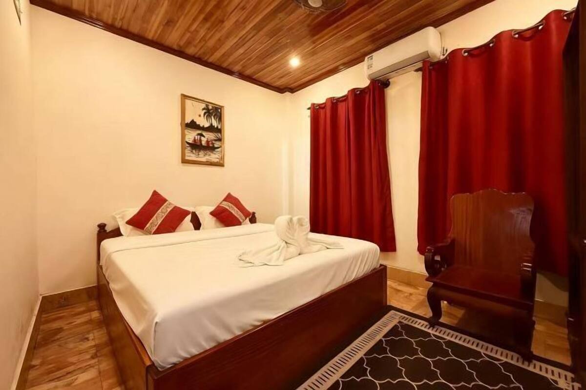 A bedroom with a wooden bed, red curtains, a wooden chair, and a painting on the wall.