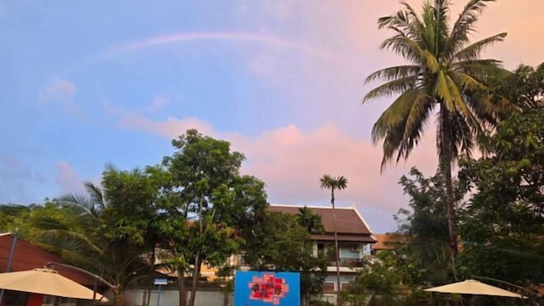 A swimming pool with a blue and orange poolside area, surrounded by palm trees and a building with a red roof. - Nan Ya Hotel(Sene Hua Phanh Guesthouse) (Luang Prabang)