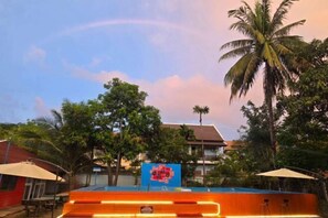 A swimming pool with a blue and orange poolside area, surrounded by palm trees and a building with a red roof. - Nan Ya Hotel(Sene Hua Phanh Guesthouse) (Luang Prabang)