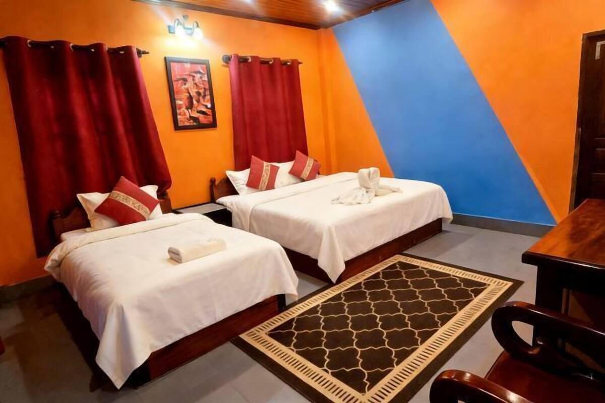 A hotel room with two beds, red curtains, a wooden table, and a patterned rug.