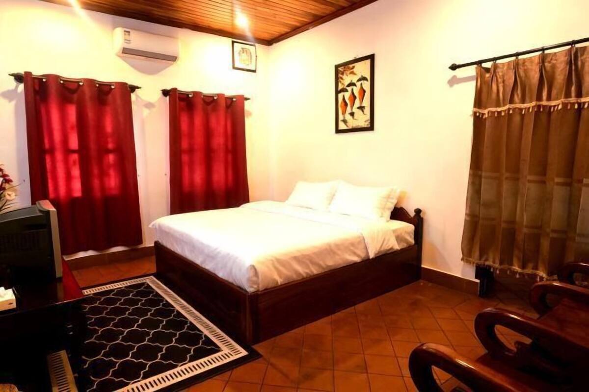 A bedroom with a bed, red curtains, a brown curtain, a framed picture on the wall, and a wooden ceiling.