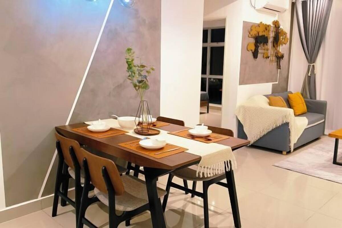 Ein Essbereich mit einem Holztisch und Stühlen, einer Vase mit grünen Blättern und einer grauen Couch mit gelben Kissen im Hintergrund.