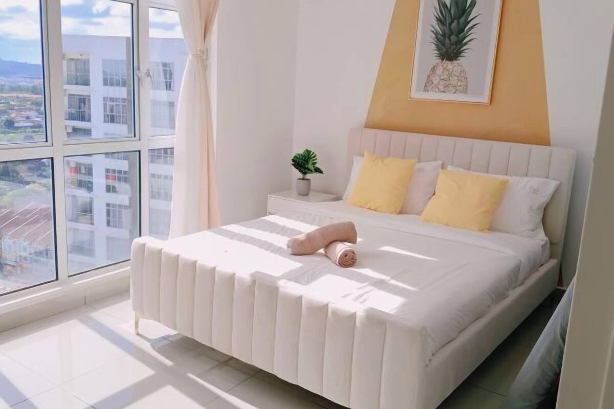 Ein Schlafzimmer mit einem Bett, einem Nachttisch mit Pflanze, einem Fenster mit Blick auf Gebäude und einer Ananas-Kunstwerk an der Wand.