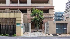 Un edificio de varios pisos con una entrada protegida por una reja, un pequeño árbol en el centro y un letrero que indica una zona de estacionamiento.
