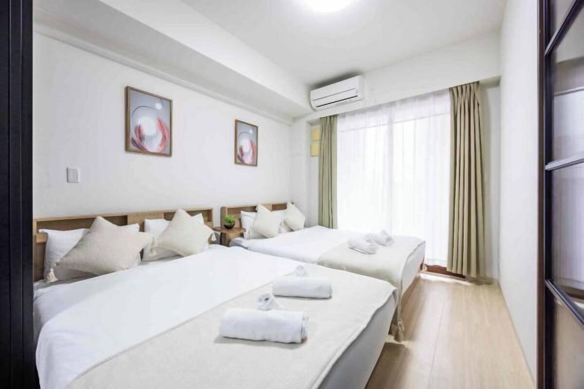 Quarto de hotel com duas camas, uma janela grande com cortinas, ar condicionado e uma obra de arte na parede.