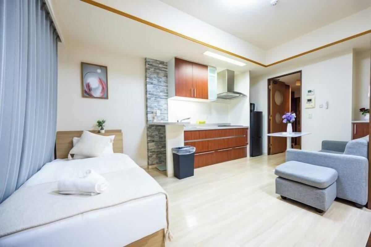 Quarto de hotel moderno com cama, pequena cozinha, área de estar e banheiro visível através de uma porta aberta.