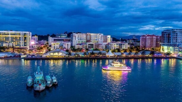 Eine Uferlandschaft mit Gebäuden, einer Marina mit Booten und einem Fluss, der die Stadtlichter spiegelt.