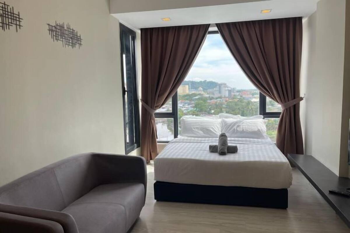 A bedroom with a bed, a sofa, and a view of the city through large windows.