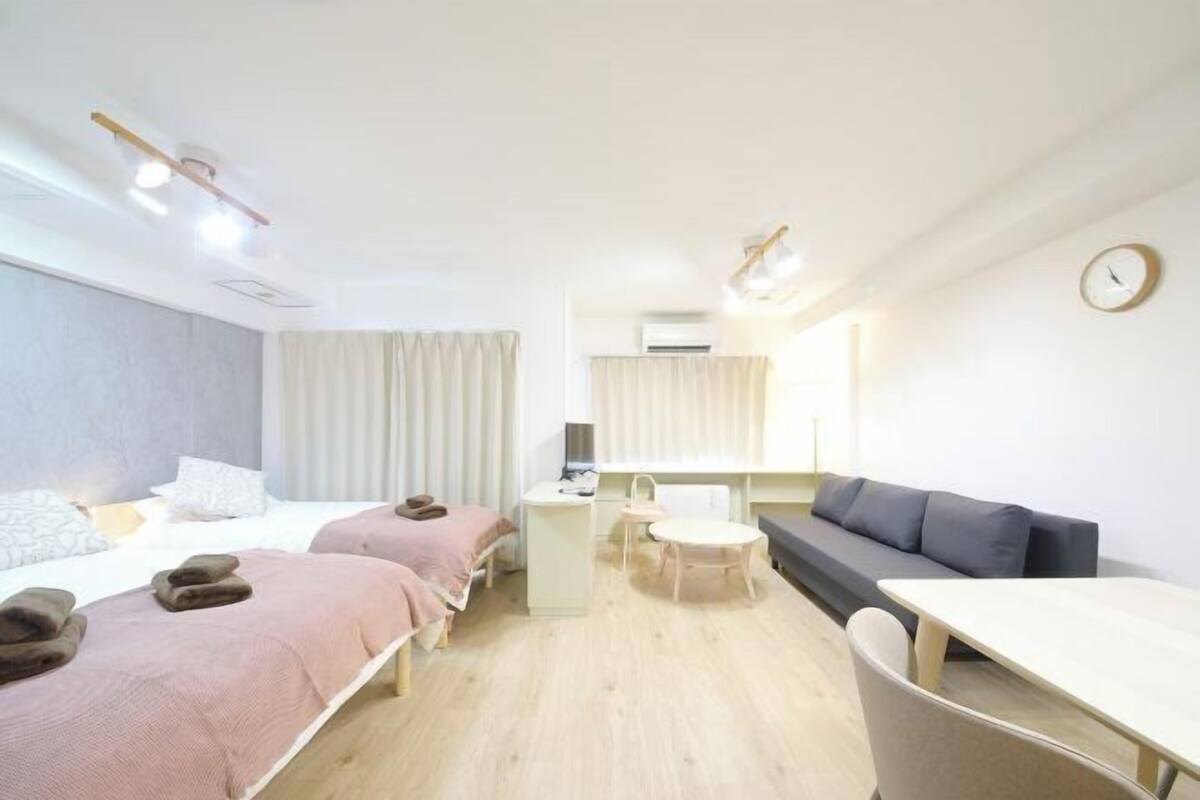 A hotel room with two beds, a desk, a chair, a sofa, a clock, and a window with curtains.