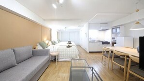 A modern, well-lit room with a grey sofa, a small round table, a wooden dining table, and a kitchenette in the background. - Northern Hills Minami 6 (Sapporo)