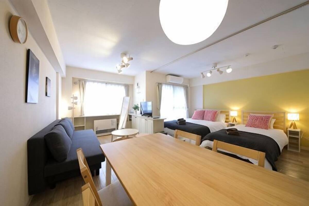 A room with a wooden dining table, two single beds, a sofa, a TV, and a desk with a chair.