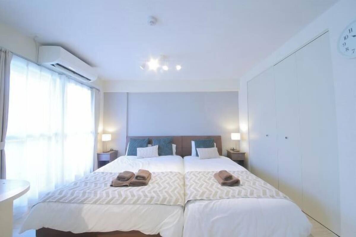 A hotel room with two beds, a large window with curtains, an air conditioning unit, a clock on the wall, and a wardrobe.