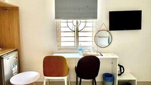 A small room with a desk, two chairs, a television, a window with a blind, a mirror, and a trash bin. - Atusy Boutique Hotel (Ho Chi Minh City)