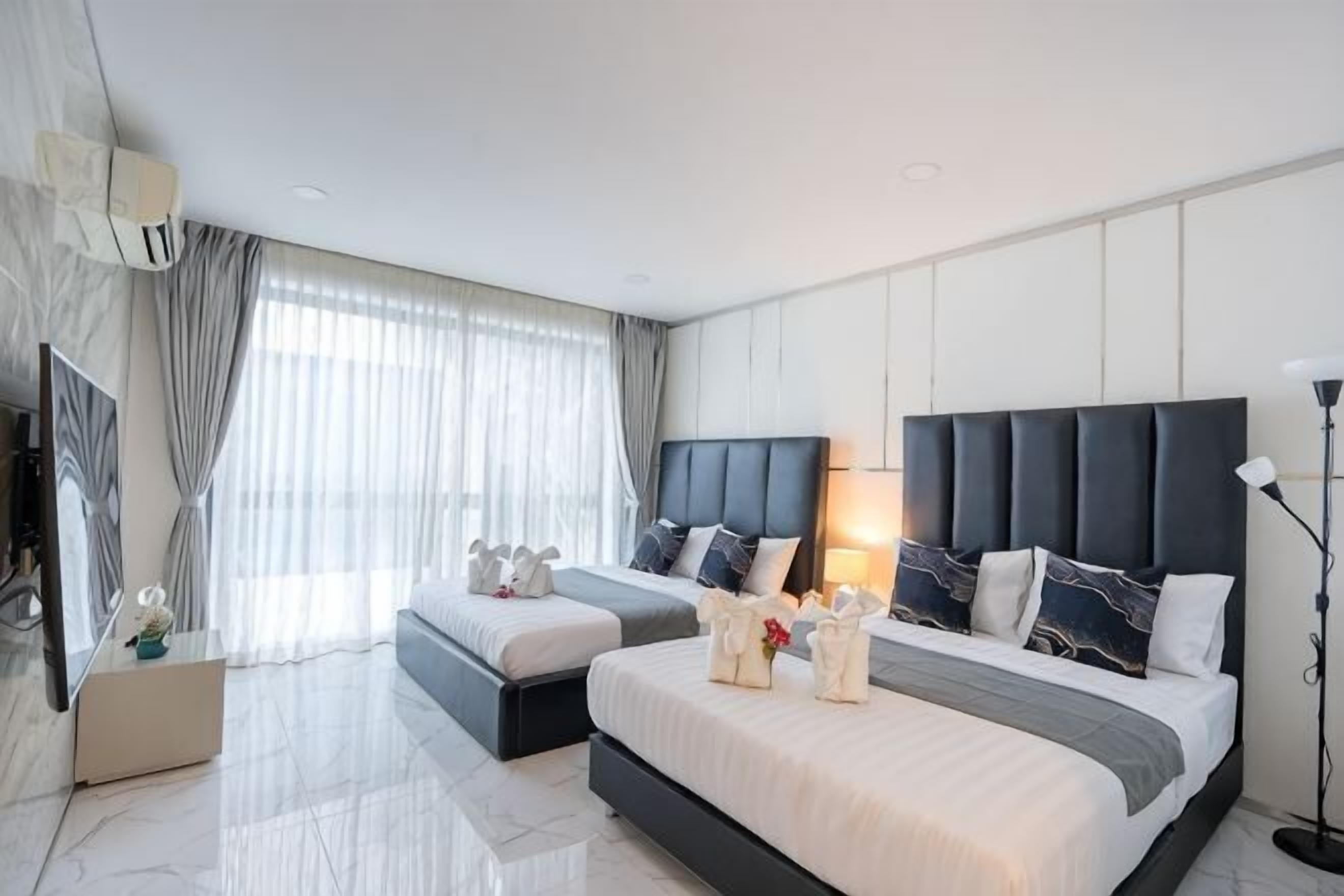 Une chambre d’hôtel moderne avec deux lits, une grande fenêtre avec des rideaux, un téléviseur à écran plat et une table de chevet avec une lampe.