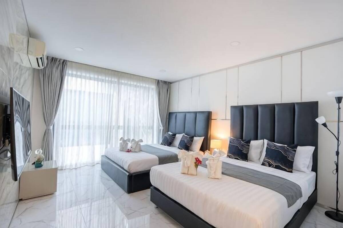 A modern hotel room with two beds, a large window with curtains, a flat-screen TV, and a bedside table with a lamp.