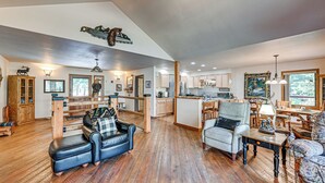 A spacious living area with a wooden floor, a black leather sofa, a grey armchair, and a dining table with chairs. A kitchen area is visible in the background with wooden cabinets and a refrigerator. - Hike, Fish & Unwind: Remote Escape in Montana (Saint Regis)