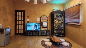 Une pièce avec un plafond en bois, un téléviseur, une armoire contenant des objets décoratifs, une porte et une fenêtre avec des stores.