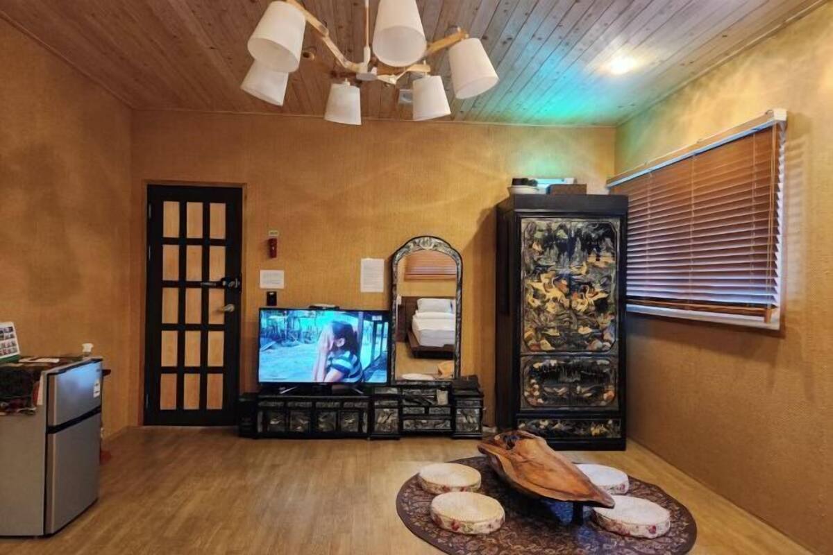 Une pièce avec un plafond en bois, un téléviseur, une armoire contenant des objets décoratifs, une porte et une fenêtre avec des stores.