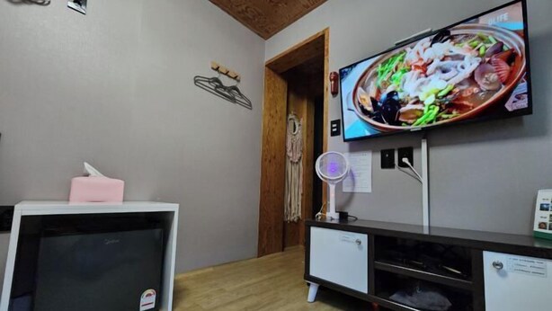 Une pièce dans laquelle est fixée un téléviseur au mur, qui diffuse une image d’un plat de fruits de mer. On y trouve également une petite armoire blanche surmontée d’une boîte de mouchoirs rose et une porte en bois.