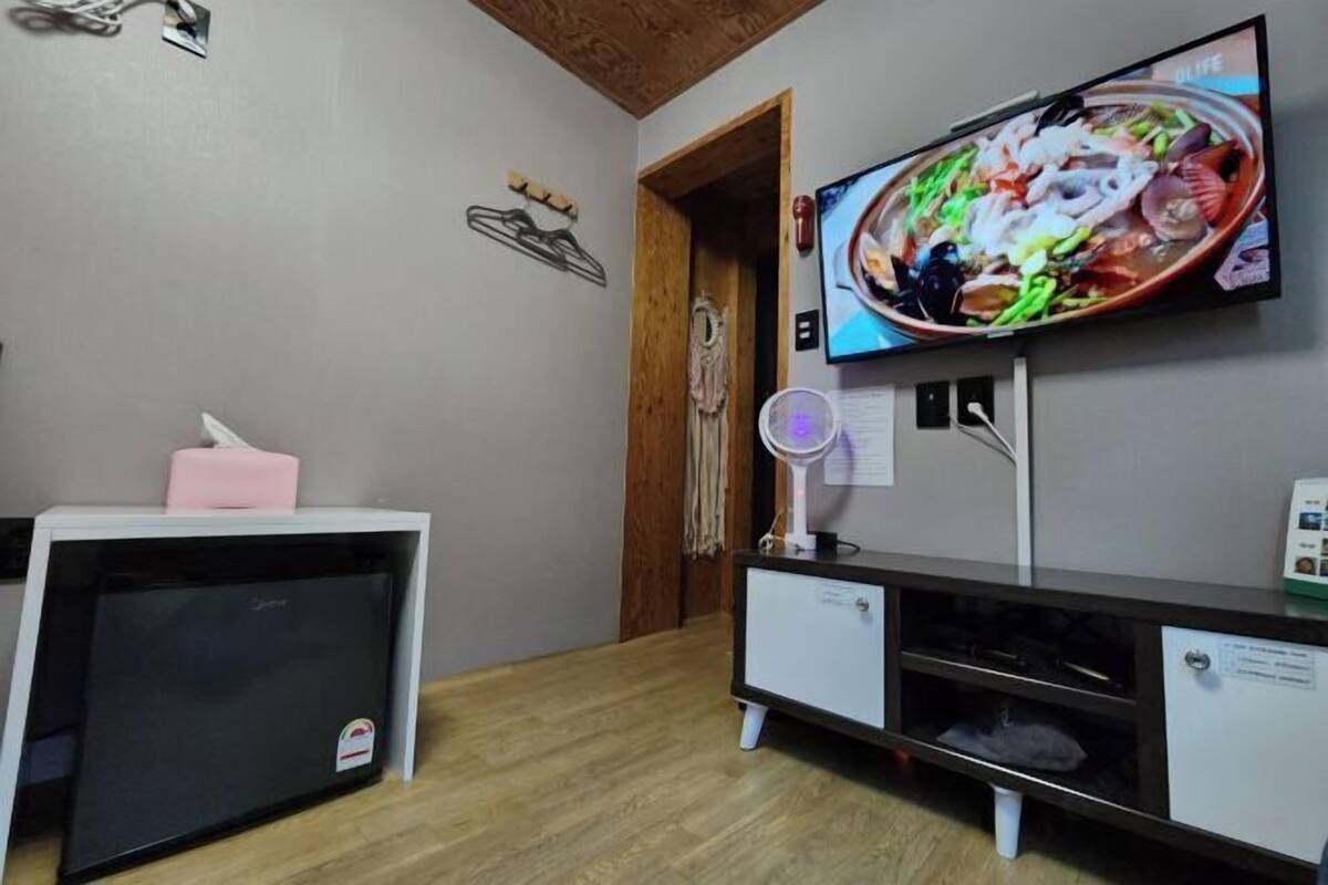 Une pièce dans laquelle est fixée un téléviseur au mur, qui diffuse une image d’un plat de fruits de mer. On y trouve également une petite armoire blanche surmontée d’une boîte de mouchoirs rose et une porte en bois.
