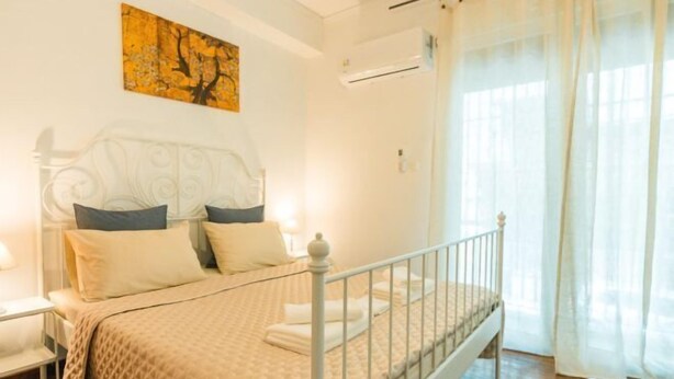 A bedroom with a bed, a nightstand, a wall-mounted air conditioner, and a window with sheer curtains. - Ma de Bright Stay Attiki Metro (Athens)