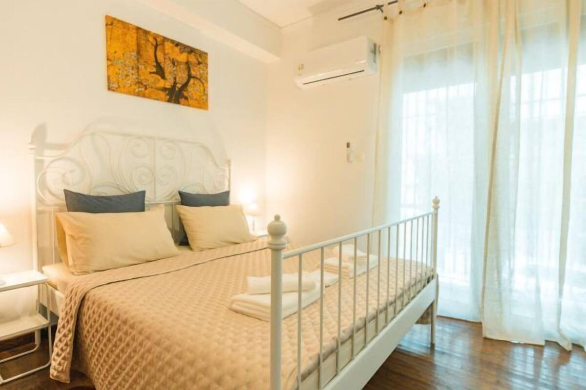 A bedroom with a bed, a nightstand, a wall-mounted air conditioner, and a window with sheer curtains.