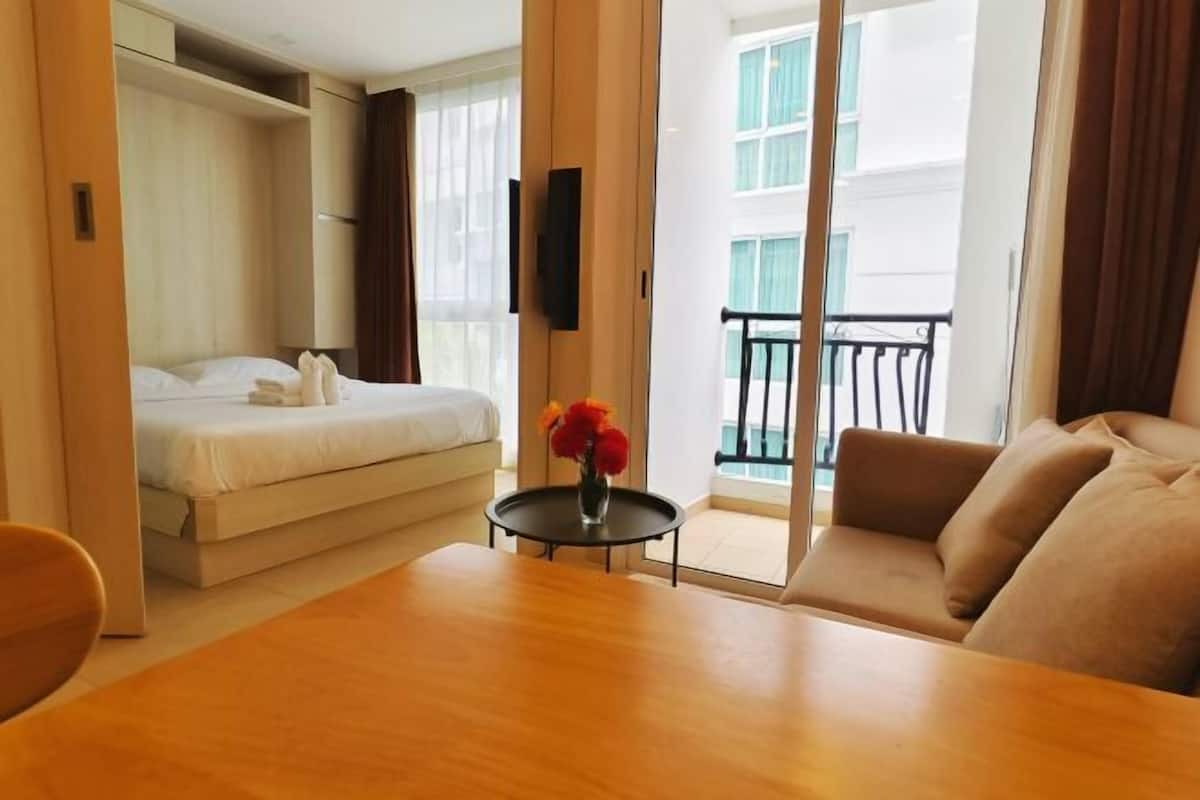 A hotel room with a bed, a sofa, a dining table, and a balcony with a railing.