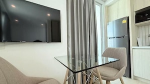 Ein zeitgemäßer Wohnbereich mit Glastisch, zwei Stühlen, einem Flachbildfernseher, einer Küchenzeile inklusive Kühlschrank und einem Fenster mit Vorhängen.