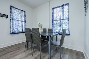 A dining area with a glass table and six chairs, two windows with blue patterned curtains, and a sign on the wall.