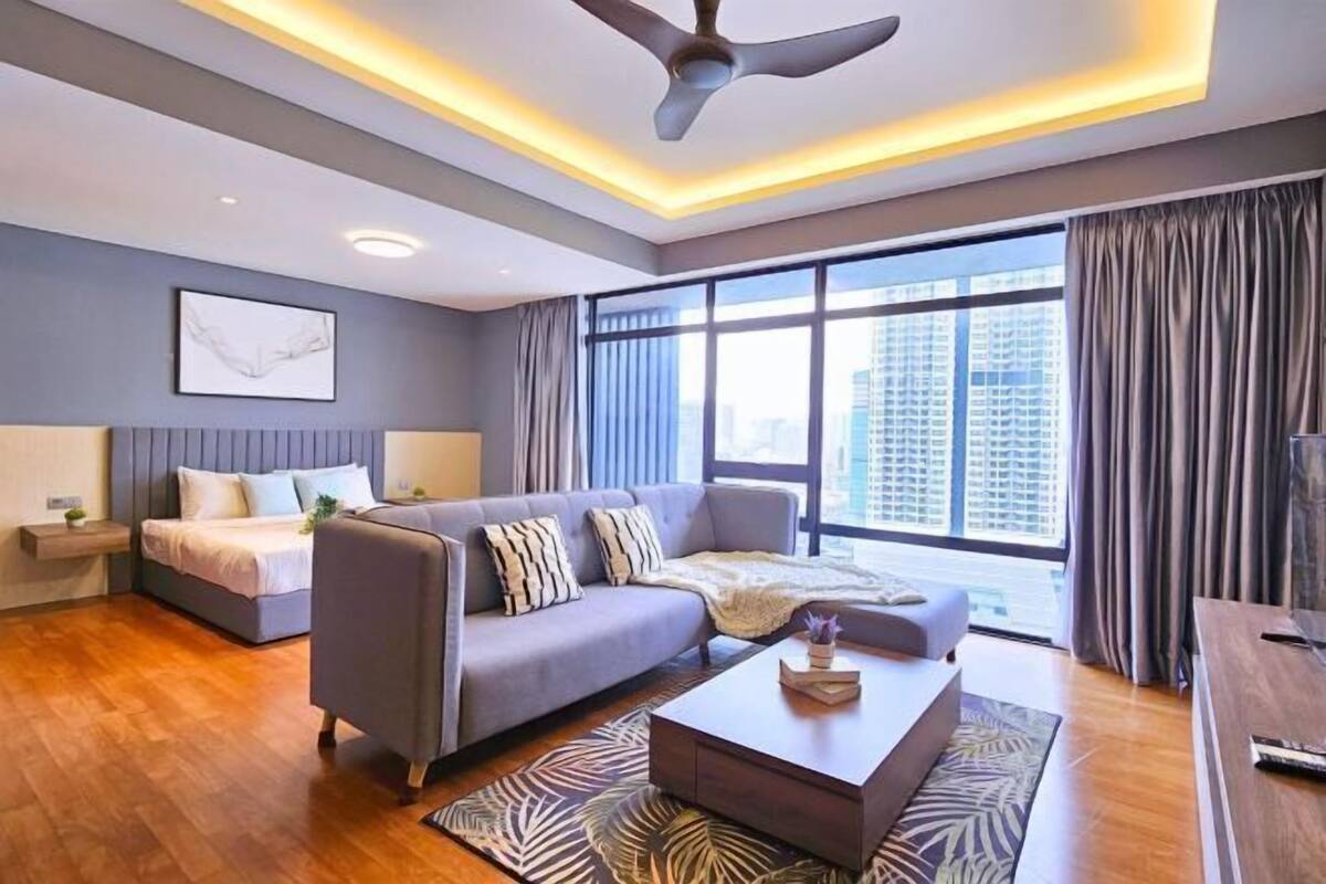 A modern hotel room with a bed, sofa, and coffee table. There is a large window with curtains and a view of buildings.