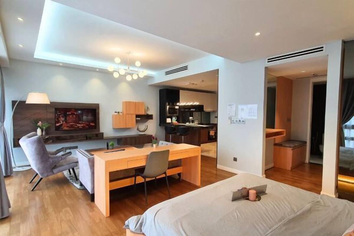 A modern hotel room with a bed, desk, chair, TV, kitchenette, and a view of the outside.