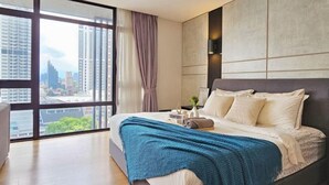 A modern bedroom with a large window, a bed with a blue blanket, a nightstand with a lamp, and a view of the cityscape. - Anggun KLCC Spacious Studio Steps to Monorail & LRT FREE Parking (Kuala Lumpur)