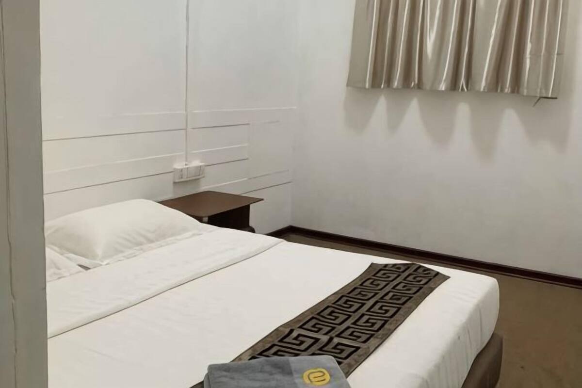 A hotel room with a bed, a nightstand, a wall-mounted air conditioning unit, and a window with curtains.