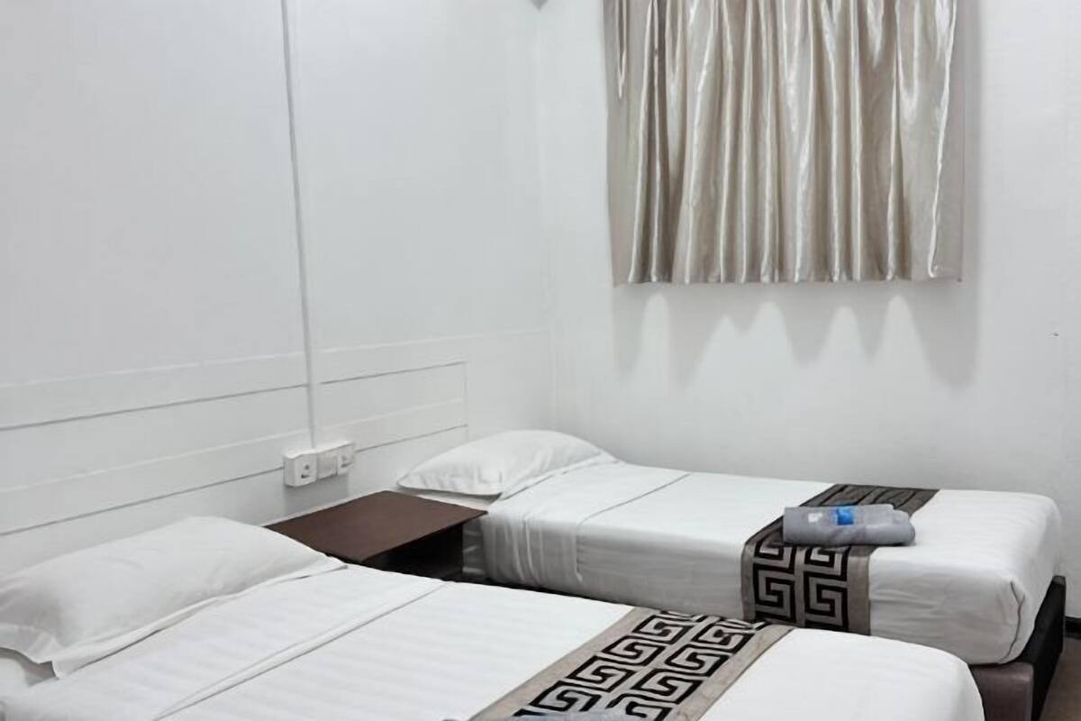 A hotel room with two single beds, a nightstand, a window with curtains, and a wall-mounted outlet.