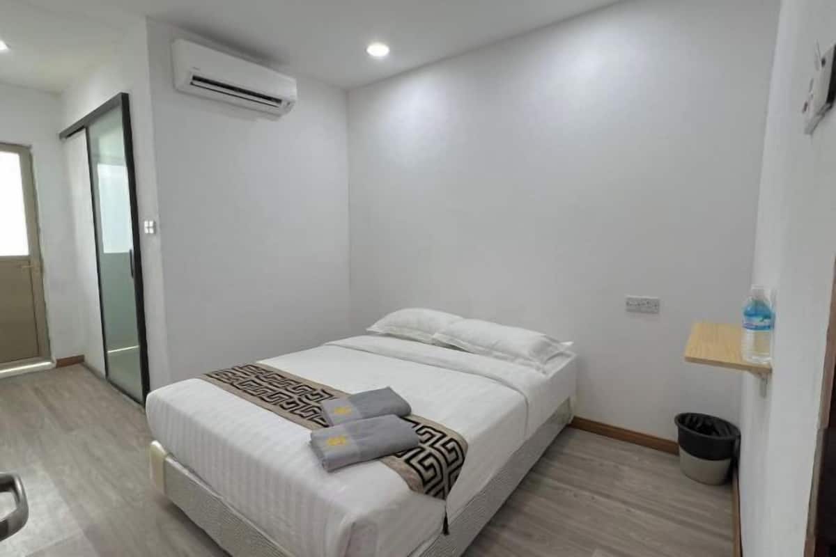 A hotel room with a bed, a small bedside table, an air conditioning unit, a trash bin, and a wooden shelf.