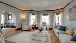 Een ruime woonkamer met een grote bank, twee fauteuils, een salontafel en een vloerkleed. Er zijn ramen met gordijnen, een lamp en een schilderij aan de muur.