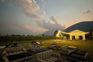 An outdoor seating area with couches and a dining table, a building with string lights, and a mountainous backdrop. - Paradise Resort & Cafe (Chiang Dao)